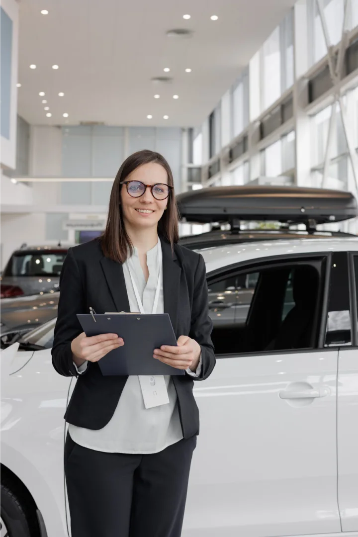 Eine lächelnde Frau in einem Anzug hält ein Clipboard in einem modernen Autohaus, bereit, potenziellen Kunden zu helfen.